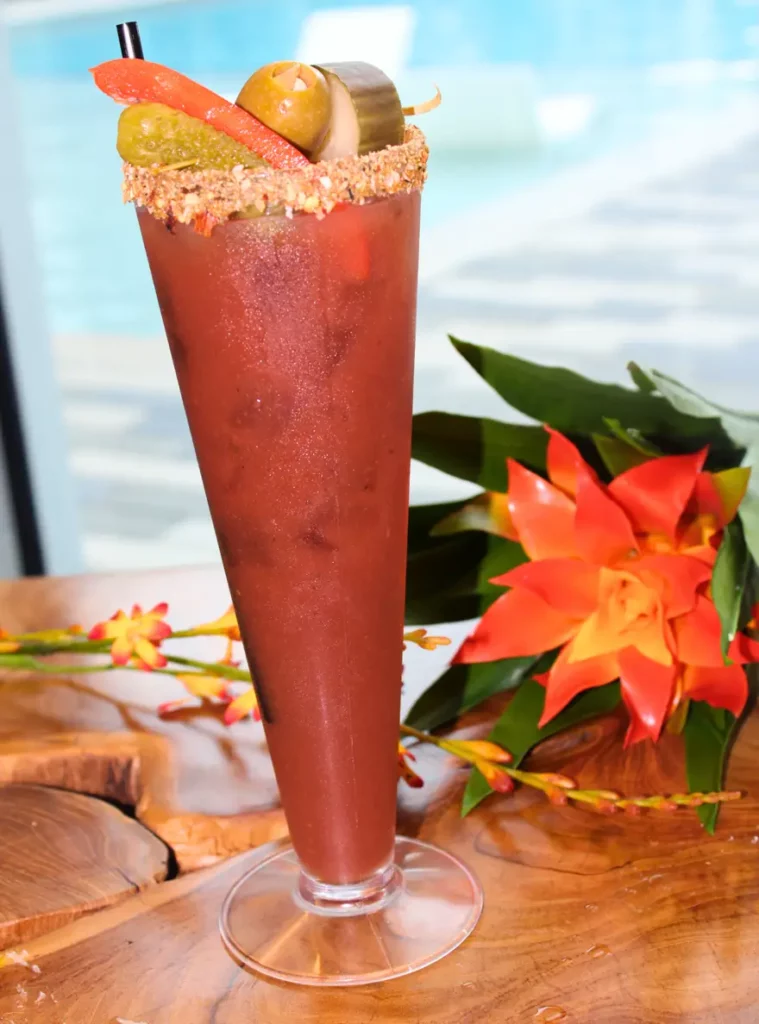 Tall glass of a Bloody Mary with garnishes, set on a wooden table near tropical flowers.