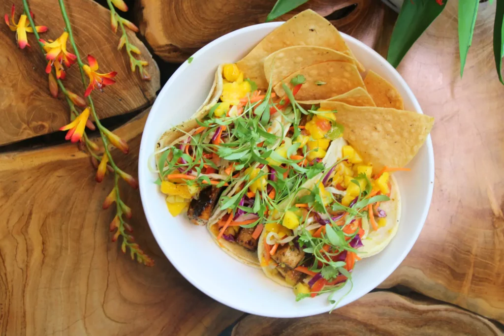 A bowl of colorful tacos with mango salsa, fresh greens, and tortilla chips on a wooden table, accompanied by vibrant orange flowers.