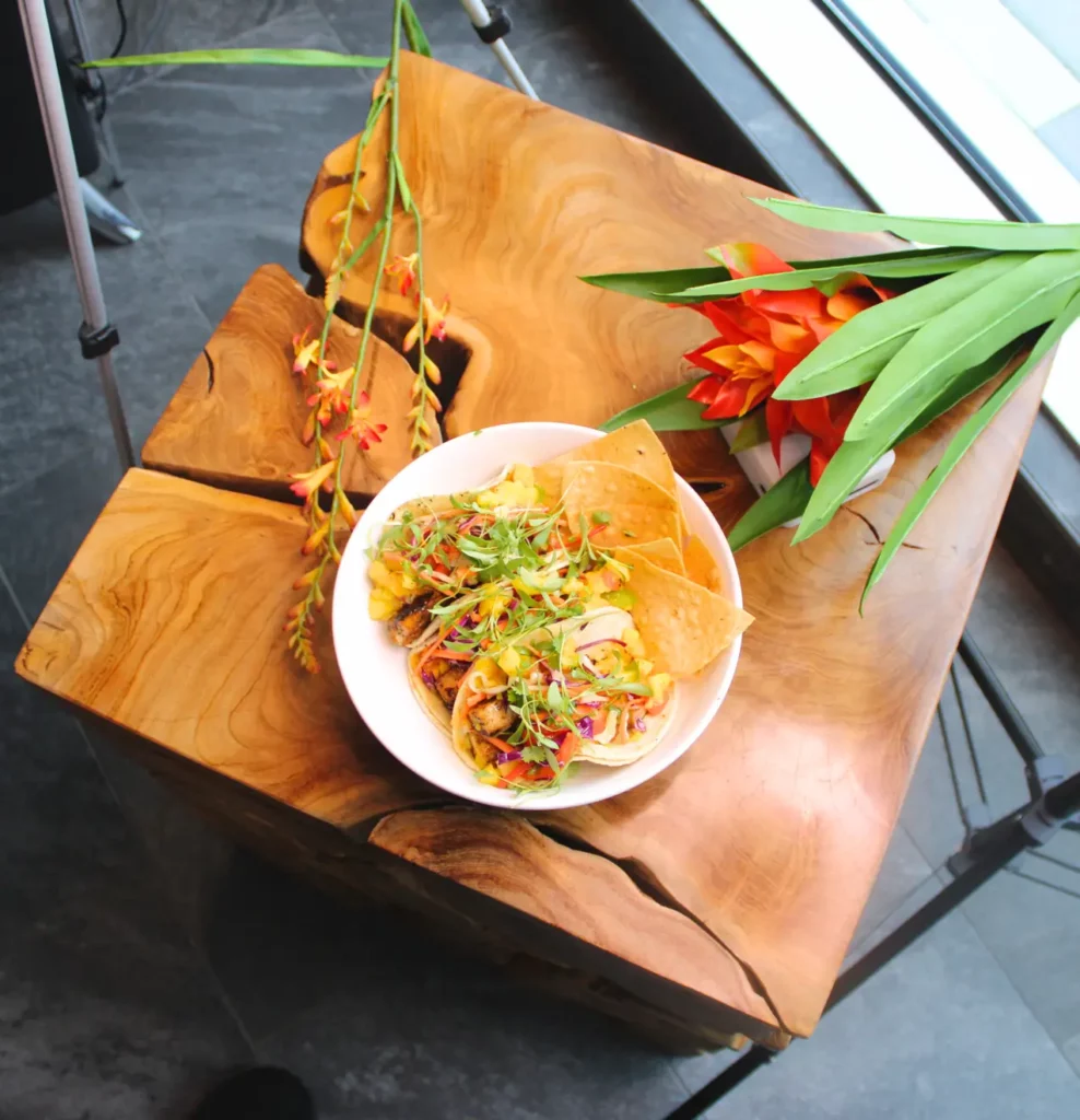A bowl of tacos with garnishes and tortilla chips on a wooden table, next to decorative flowers.