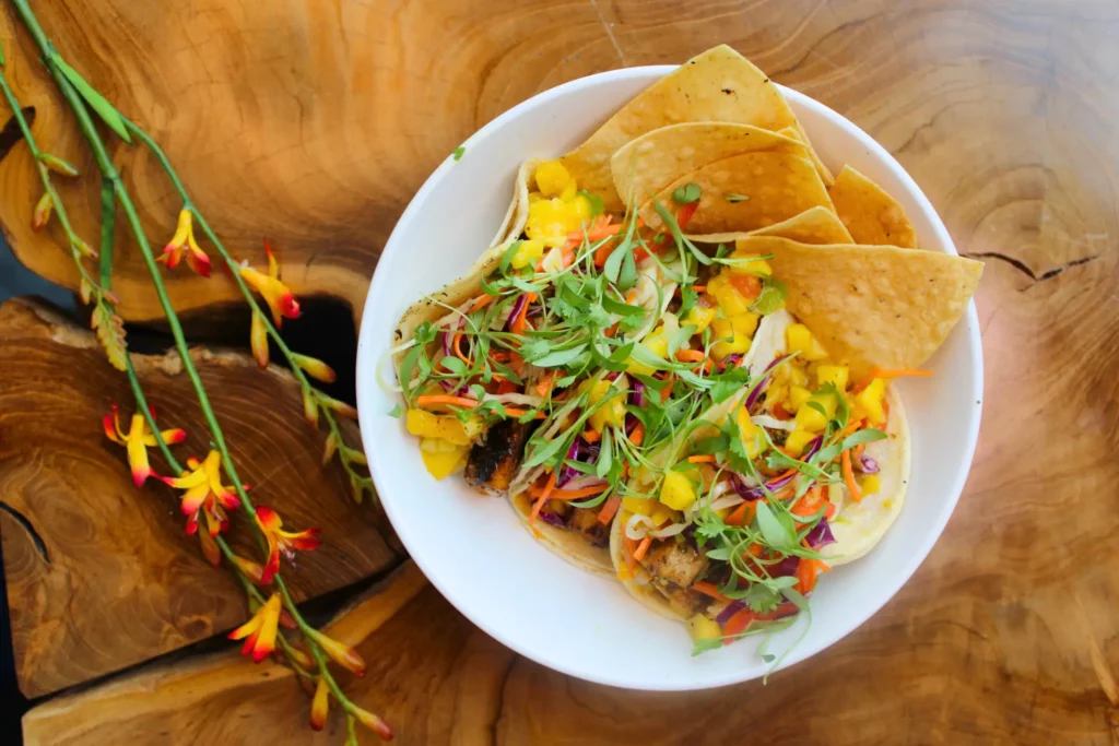 Bowl of colorful tacos garnished with greens, served with tortilla chips on a wooden table.
