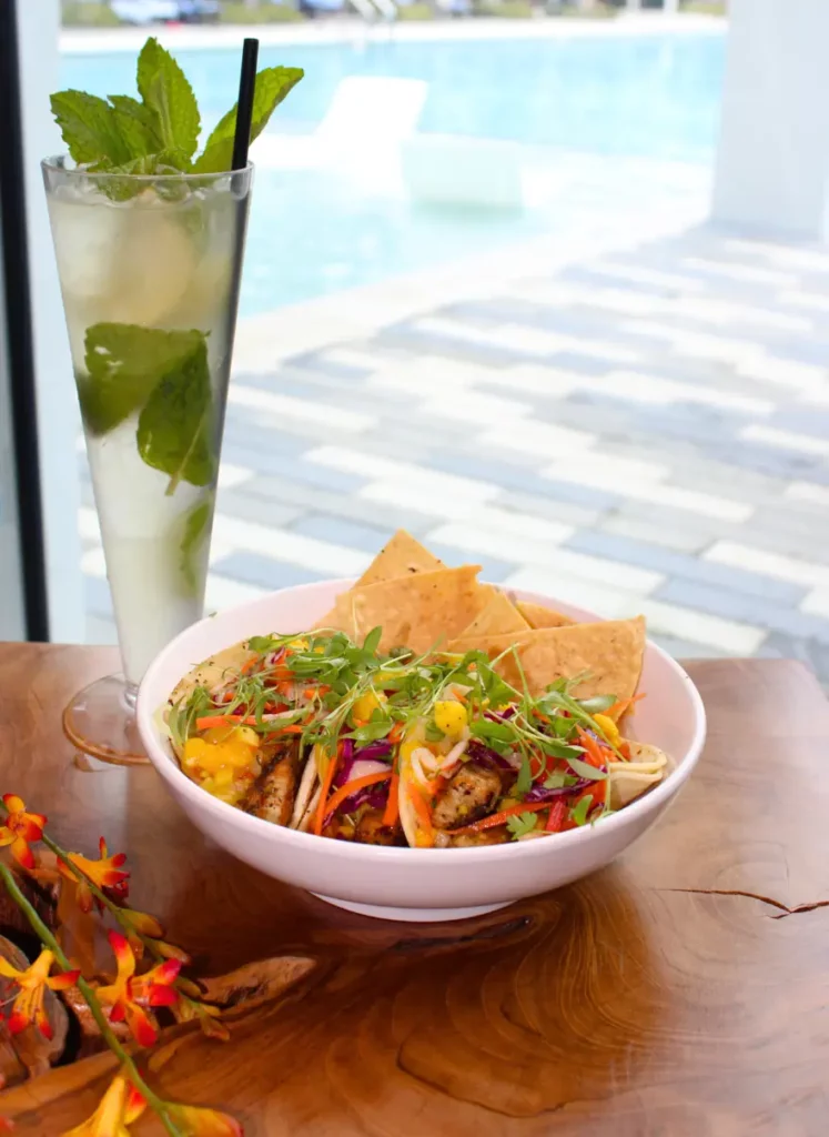 Bowl of colorful salad with chips next to a tall iced drink with mint leaves on a wooden table by a pool.