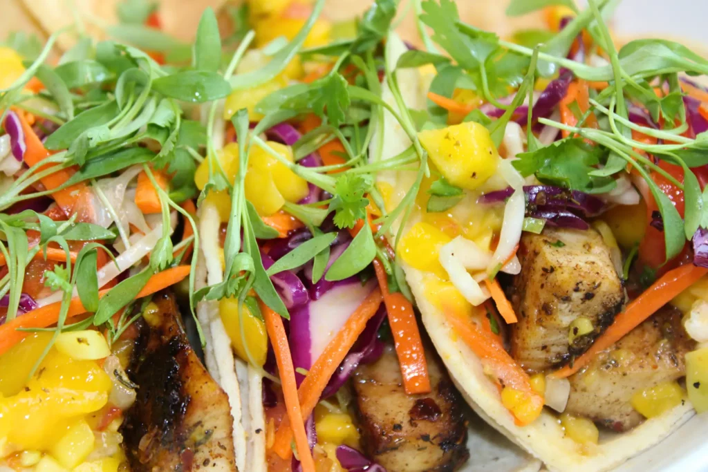 Close-up of colorful tacos filled with grilled fish, mango salsa, cabbage, carrots, and fresh greens.