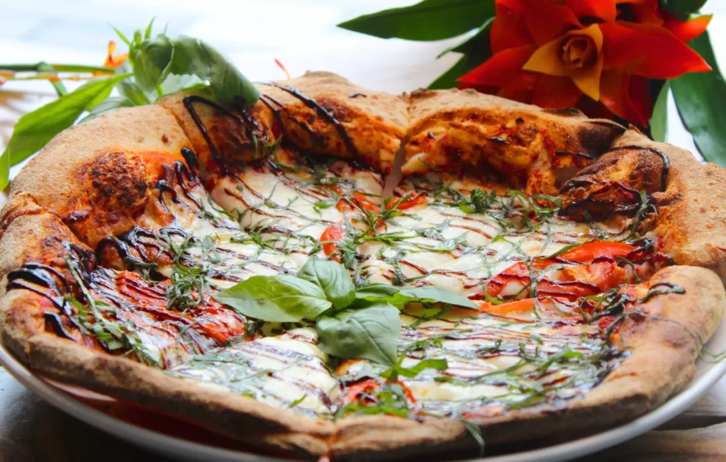 A freshly baked pizza topped with mozzarella, tomatoes, basil, and a drizzle of balsamic glaze, served on a white plate and garnished with fresh basil leaves.