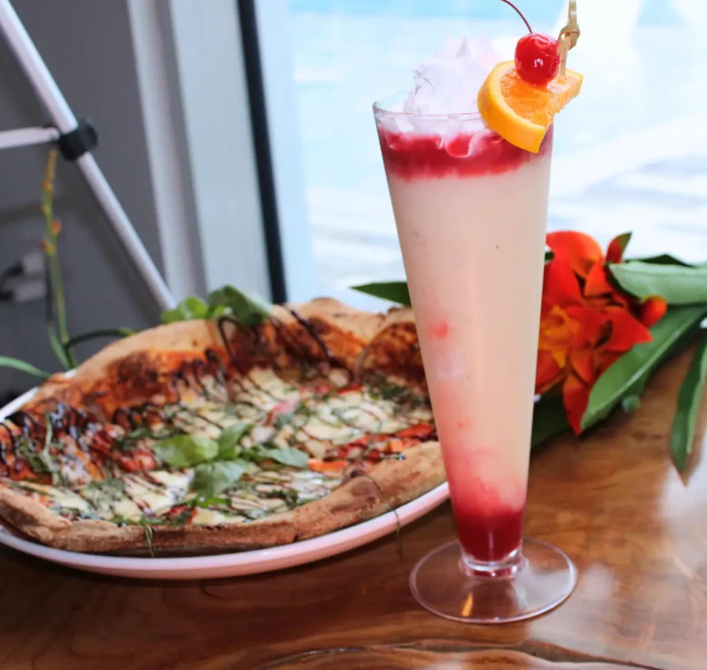 Tall glass of a layered drink with a cherry and orange slice garnish, next to a pizza on a wooden table with orange flowers in the background.