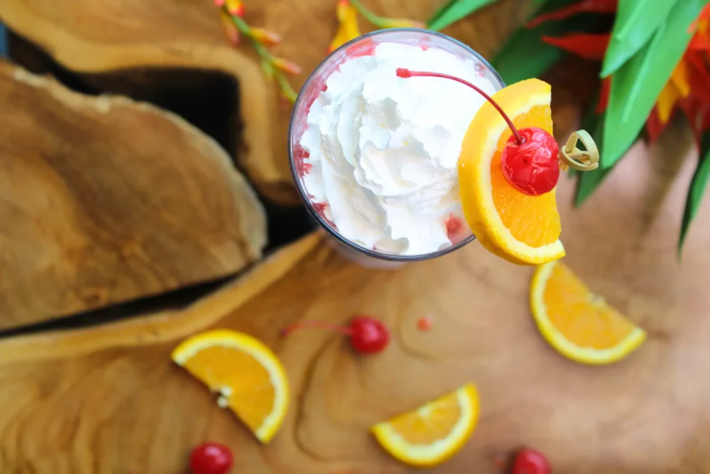 A drink topped with whipped cream, garnished with a cherry and orange slice, on a wooden surface with scattered cherries and orange slices.