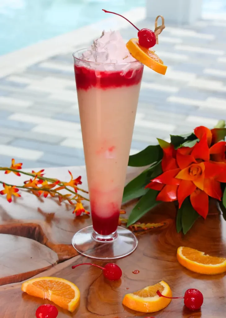 Tall glass of a creamy drink with red syrup, topped with whipped cream, cherry, and orange slice, surrounded by fruit and flowers.