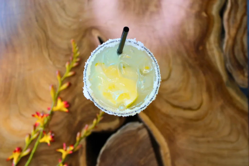 Top view of a margarita with a salted rim and straw on a wooden table, with flowers in the background.