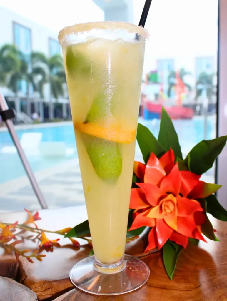 Tall glass of a tropical beverage with lime and orange slices, garnished with a sugar rim and a straw, set on a table with vibrant red flowers, poolside background.